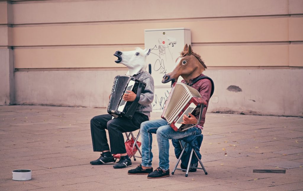 two person wearing horse heads sitting on folding chairs while playing accordions beside brown concrete building